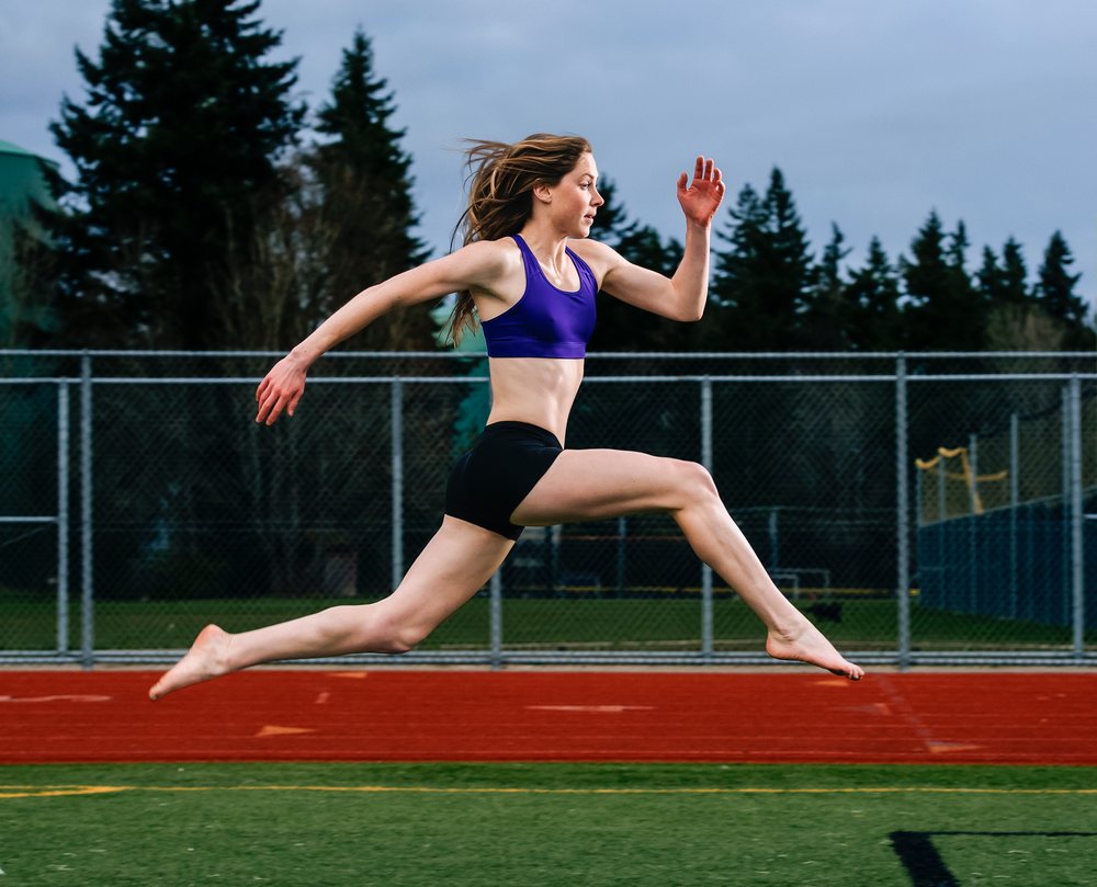Woman athlete jumping mid-air on an outdoor red running lane track field in USA during training.