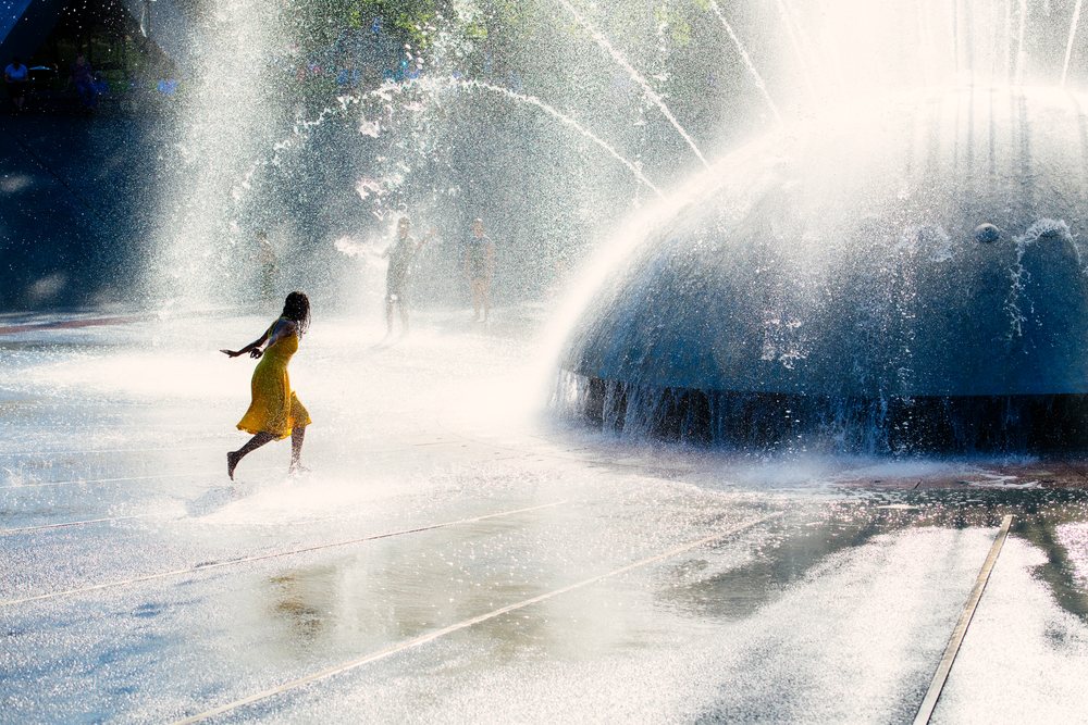 Woman running and playing in the water spray of a large outdoor fountain in a public park in the USA on a sunny day.