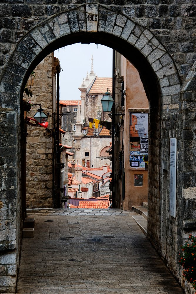 Stone archway dated 1908 frames a narrow stone-paved street lined with historic buildings.
