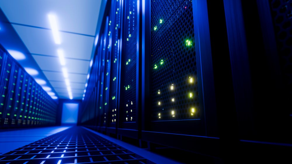 Multiple server racks with illuminated green and yellow indicator lights line a blue-lit data center corridor with vented panels and metal grid flooring.