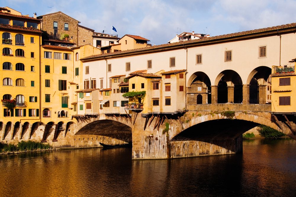 Sunlit view of Ponte Vecchio historic stone bridge with arches and multi-story buildings over a river, Florence, Italy.