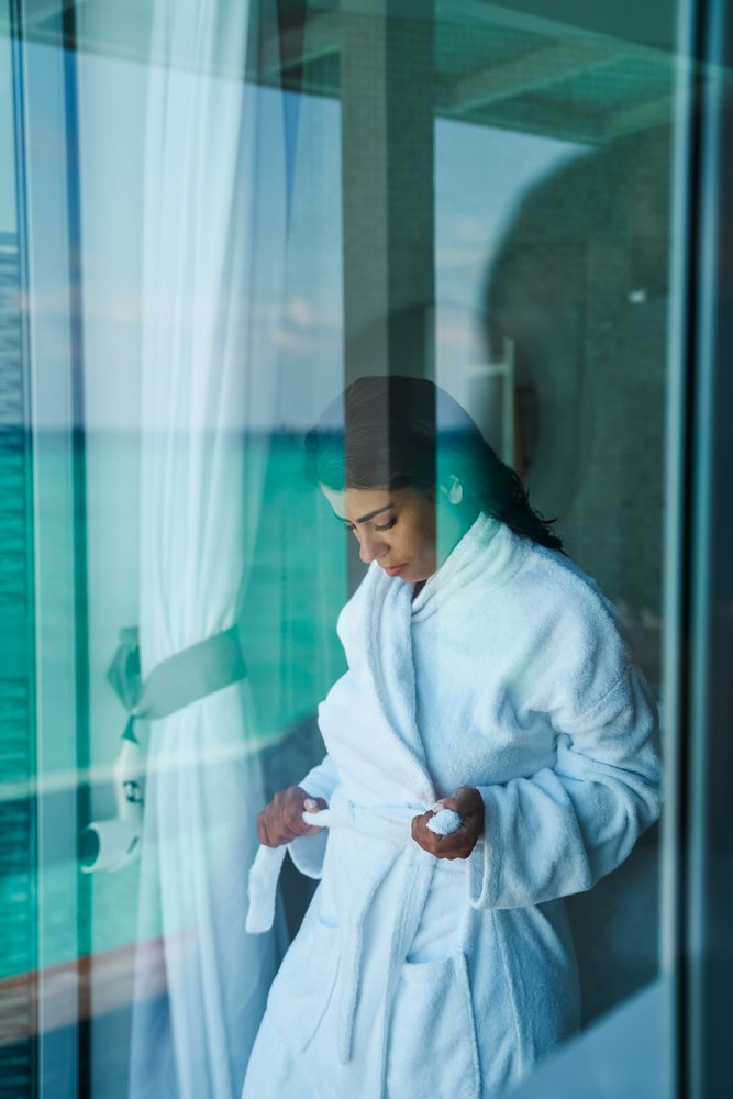 A woman in a white bathrobe ties its belt while standing indoors near a window with a view of the sea in the, Maldives.