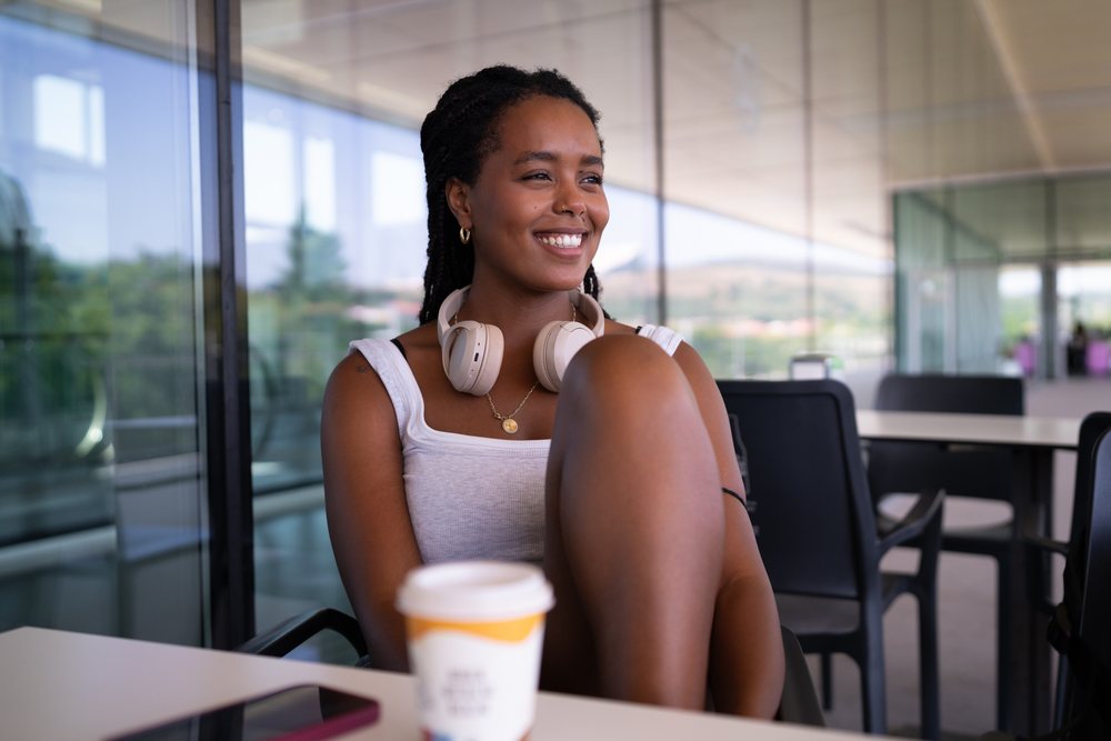 A smiling woman with headphones around her neck sits casually at a table with a coffee cup on a modern outdoor terrace, remote work, travel, Santiago De Compostela, Spain.