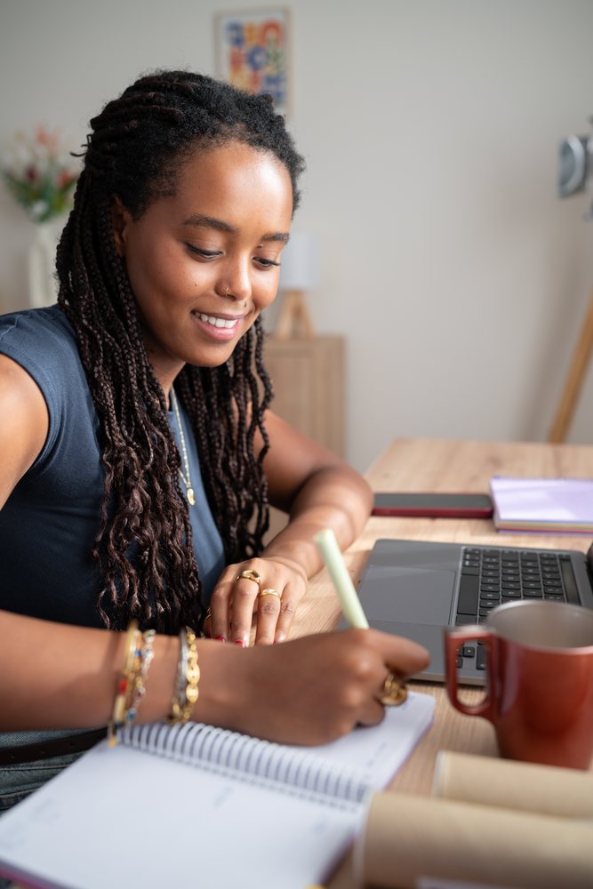 A woman with braided hair smiles while writing in a notebook at a desk in a home office, remote work, student, Spain.