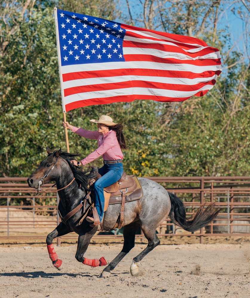 Female rider gallops a horse wearing red leg wraps while carrying a large American flag in an outdoor rodeo arena, Southwest USA.