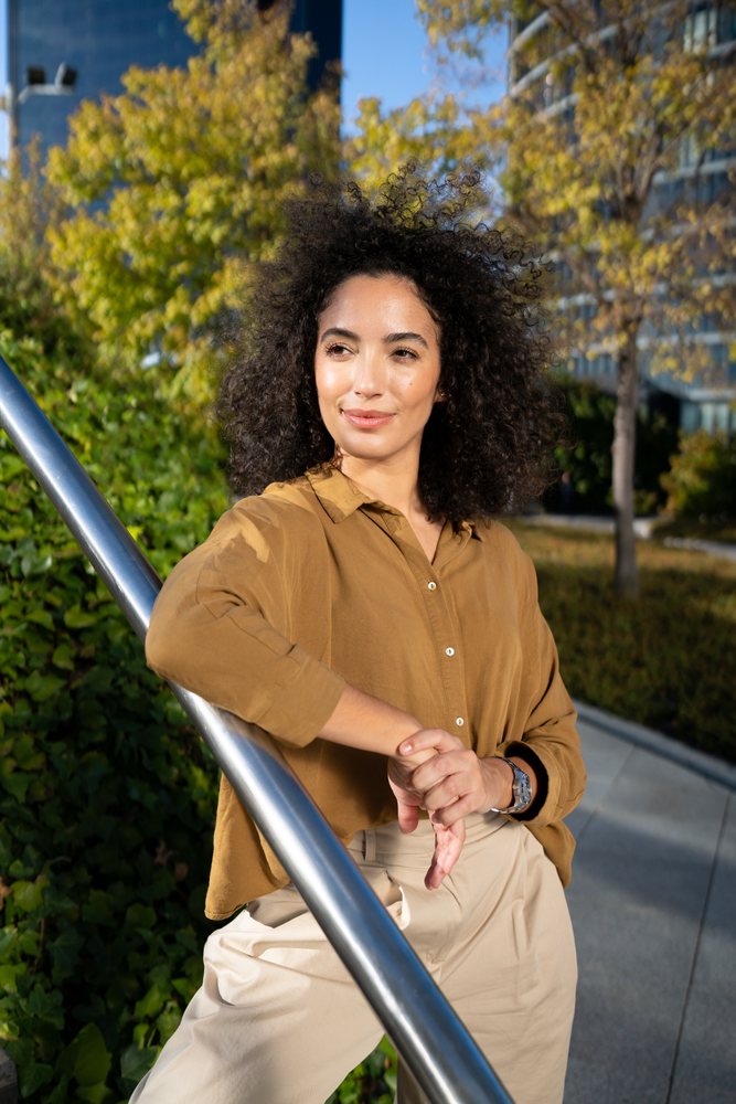 Woman with dark curly hair leaning on a metal handrail outdoors in an urban park with autumn trees and paved walkway in daylight, Madrid, Spain.