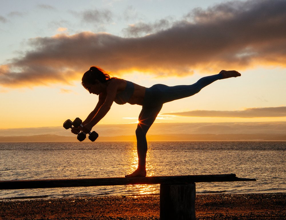 Silhouette of a woman balancing on a narrow wooden beam while holding dumbbells during sunset at a beach, WA, USA.
