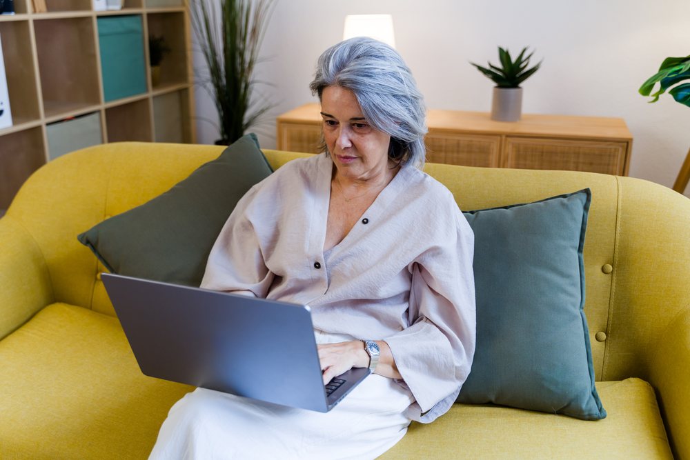 Older woman with gray hair sits on a yellow couch using a laptop indoors with green pillows and houseplants nearby, Spain.