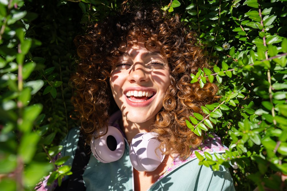 Smiling woman with curly hair and headphones outdoors, sunlight casting leaf shadows on her face amid green foliage, Spain.