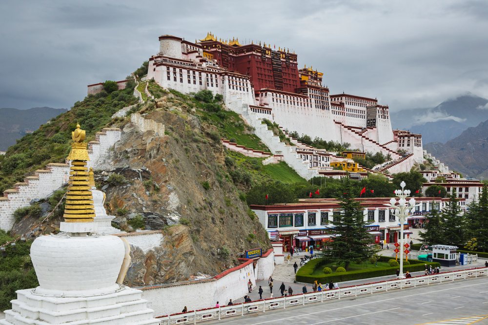 Potala Palace stands on a hill overlooking Lhasa with tourists, a white shrine, and mountainous terrain under a cloudy sky, Autonomous Region of Tibet.