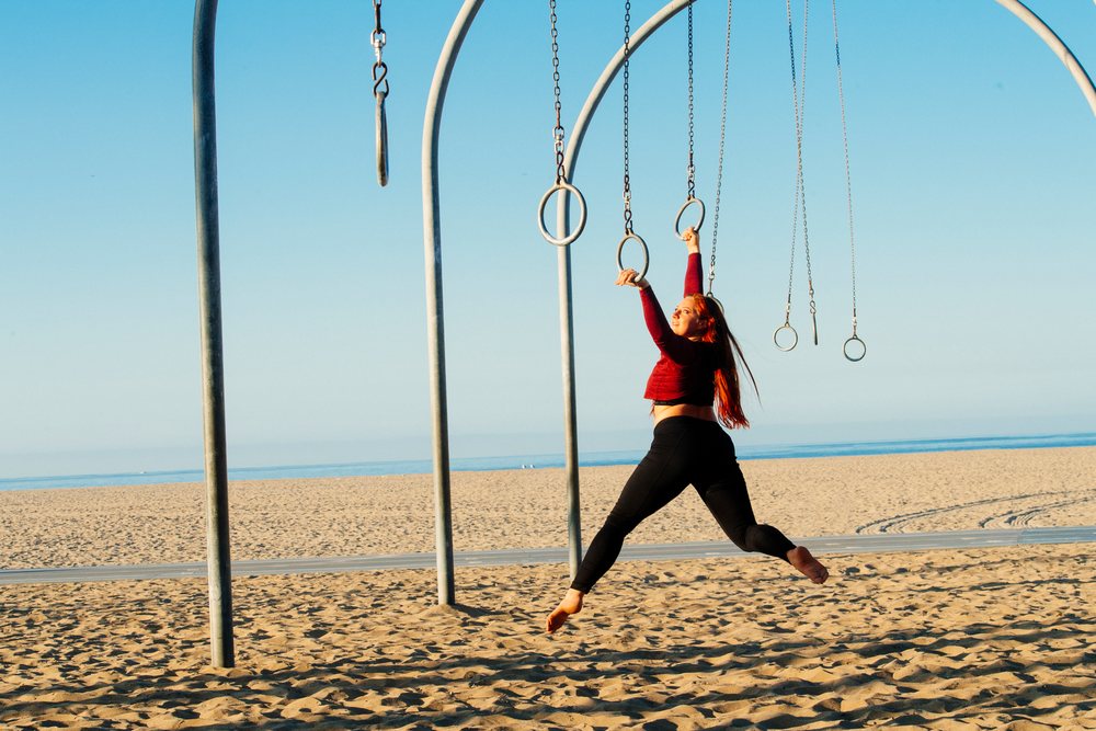 Woman holding gymnastic rings on a sandy beach with ocean and clear blue sky, California, USA.