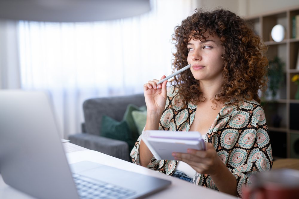Woman with curly hair sitting indoors near a laptop, holding a notebook and pen, looking thoughtfully aside, home, girl, Spain.