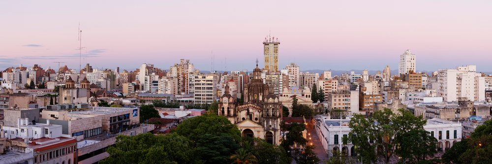 Cityscape of Cordoba, Argentina at dusk showing buildings and a cathedral under a pink sky.