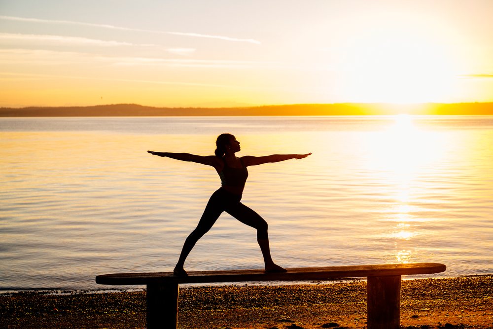 Woman practicing a yoga warrior pose on a bench by a peaceful beach at sunset, USA.