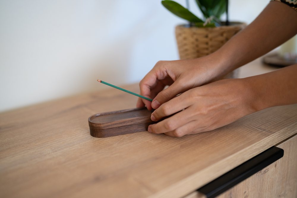 Two hands placing a green incense stick into a wooden holder on a wooden table next to a potted plant indoors, home, girl, Spain.