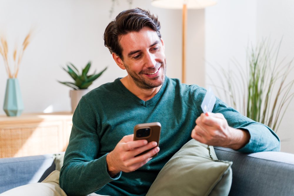 Adult man wearing a green sweater smiles while holding a smartphone and credit card sitting on a grey sofa in a daylight-lit living room, Spain.