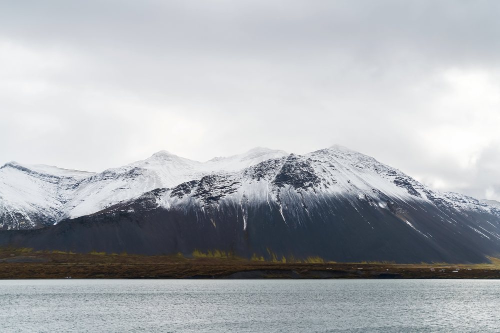 Snowy mountains beside calm water under an overcast sky, Iceland.