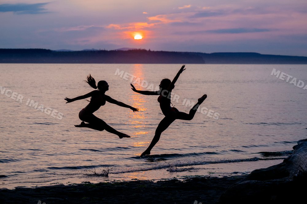 Two women perform partner acrobatics jumping on the beach at sunset