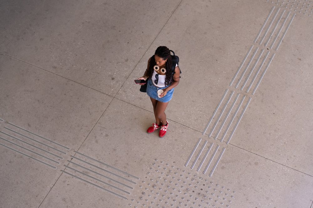 A woman stands on tactile concrete pavement holding a coffee cup and looking at her phone, remote work, travel, Santiago De Compostela, Spain.