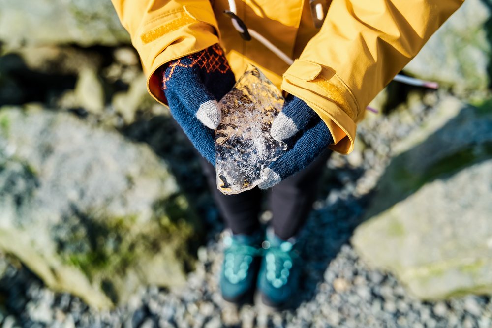 Person wearing a yellow jacket and blue gloves holding a chunk of ice outdoors on rocky ground, Iceland.