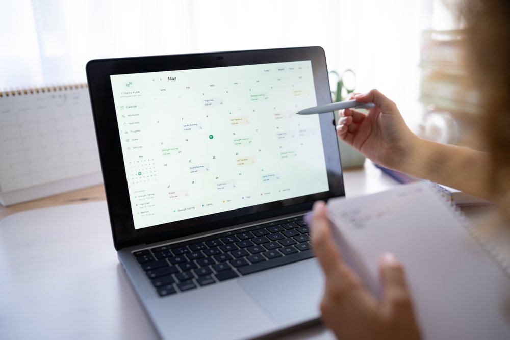 Woman holding a pen and notebook points at a laptop screen displaying a calendar with a workout schedule in a home office, Spain.