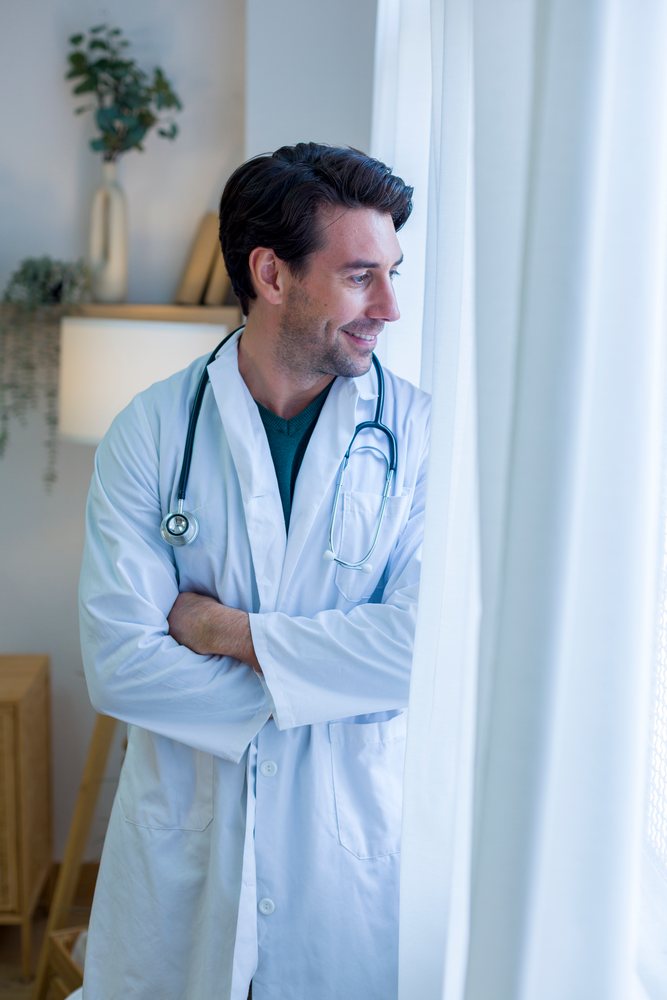 Doctor wearing white lab coat and stethoscope looks out a window inside a well-lit indoor home room with white curtains, technology, young adult.