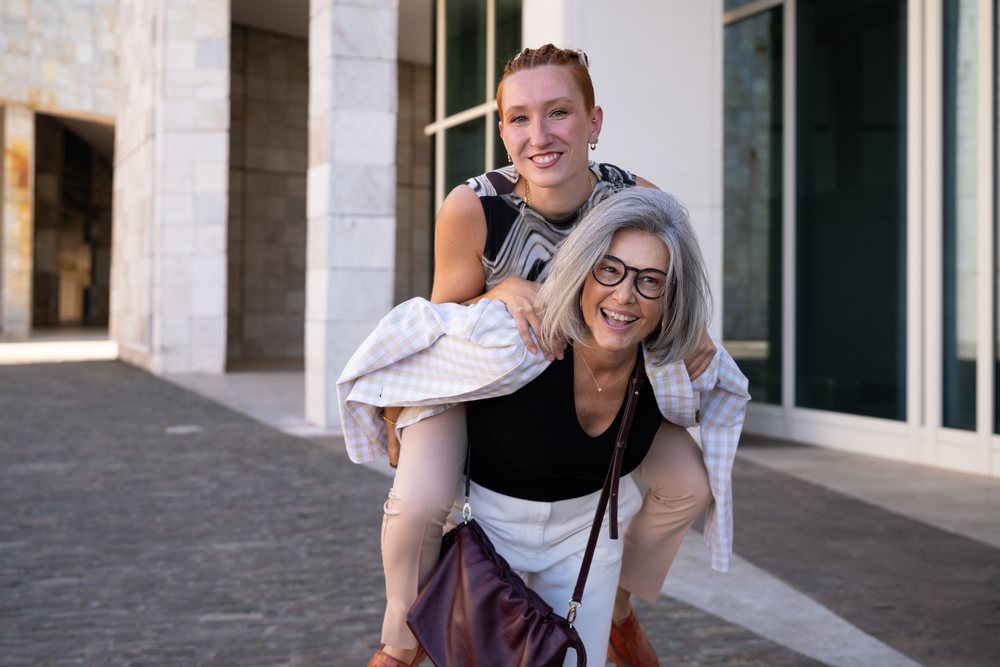 Two women sharing a joyful piggyback ride outdoors near a modern urban building, expressing friendship and happiness, businesswomen, Spain.