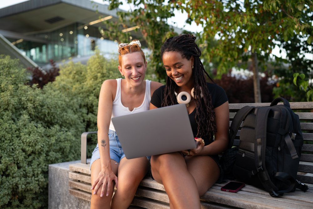 Two young women sit on a bench outdoors using a laptop together in a green urban park setting, Santiago De Compostela, Spain.