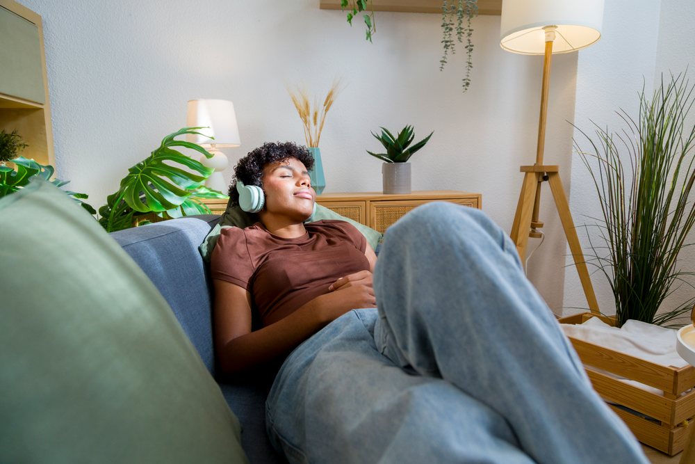 Young woman sitting on a sofa wearing headphones in a modern living room with decorative houseplants and wooden furniture.