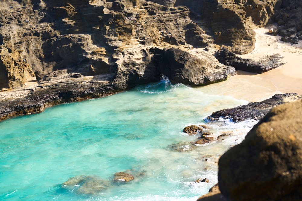 Rocky cliffs and small sandy beach meet clear blue ocean waves on the shore of Oahu, Oahu, Hawaii, USA.