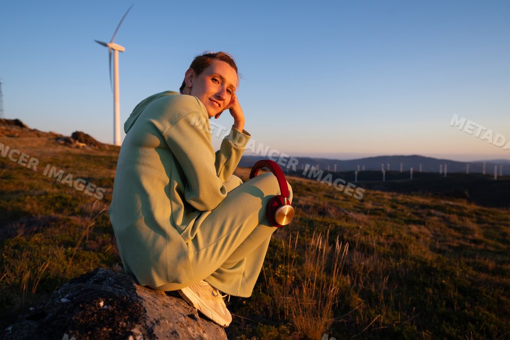 Young white woman sitting with headphones near wind turbines at sunset