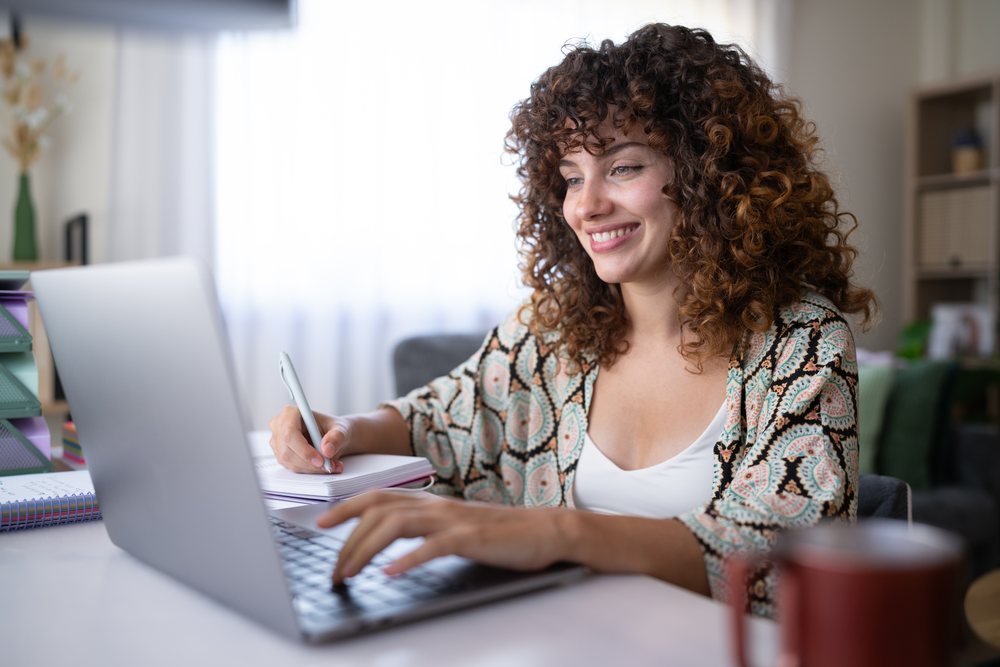Woman with curly hair smiling while writing notes and working on a laptop at a desk in a home setting, Spain.