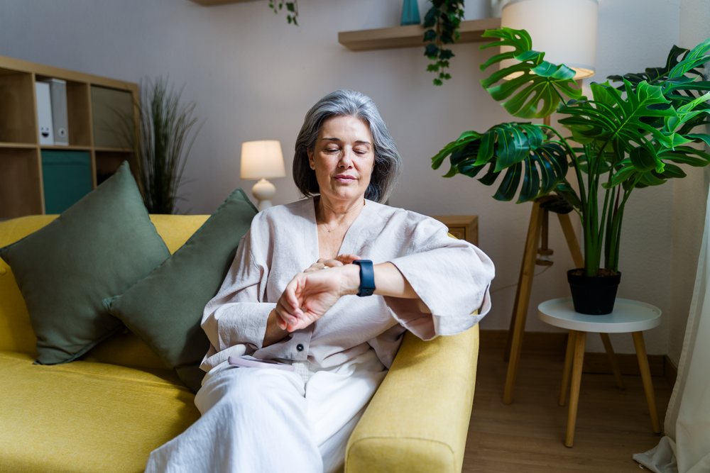 Gray-haired woman sits on a yellow sofa in a living room, checking her smart watch while relaxing, Spain.
