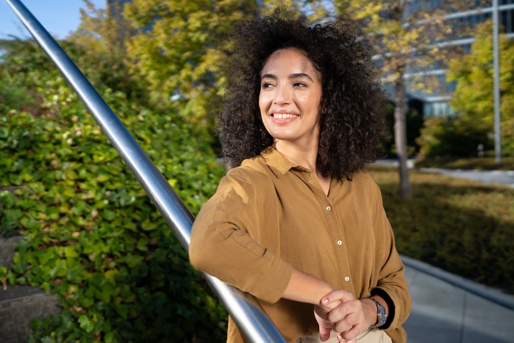 Woman with curly hair smiles leaning on a handrail in an urban park with green foliage under sunlight, Madrid, Spain.
