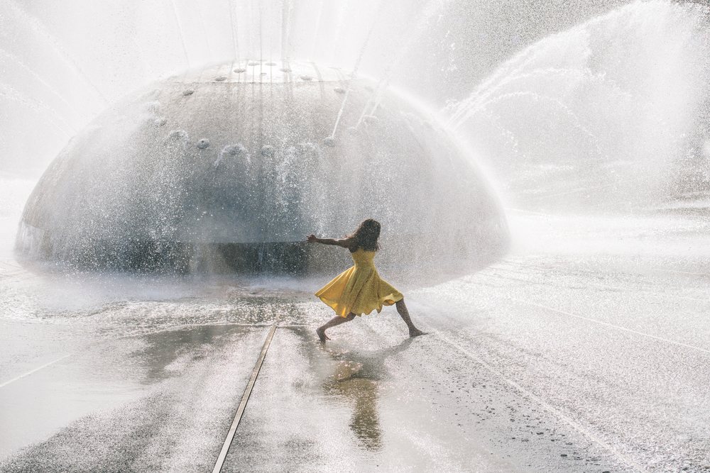 Barefoot girl wearing a yellow dress dances joyfully in the spray of an outdoor water fountain on a wet surface under sunlight, USA.
