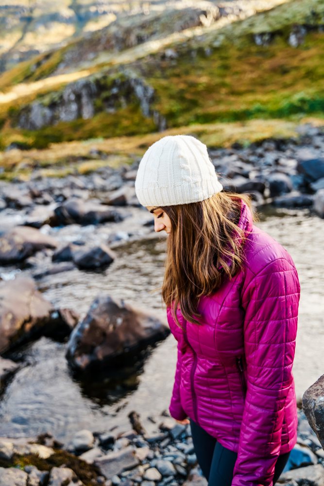 A woman wearing a red jacket and white knit hat hikes near a rocky river, Iceland.