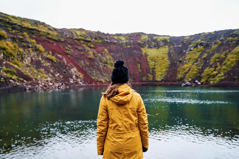 A woman wearing a yellow jacket and black knit hat stands facing a volcanic crater lake surrounded by rocky hills with moss and vegetation, Iceland.