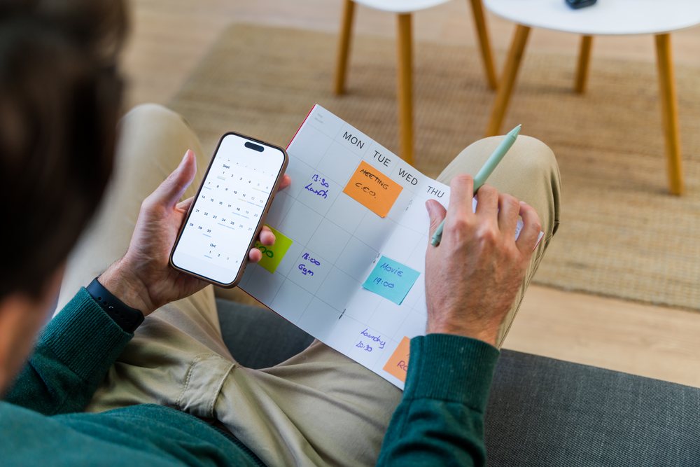 Male sits on a couch planning his monthly schedule with a calendar planner and smartphone in a home setting, Spain.