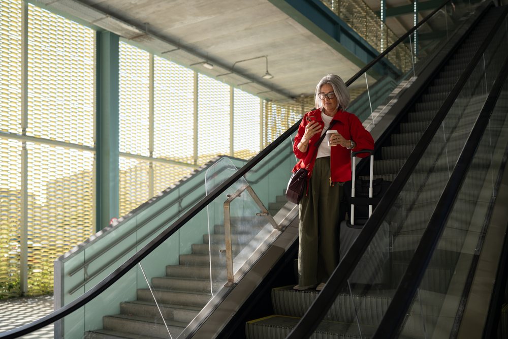 Middle-aged woman with gray hair holding coffee and smartphone stands on an indoor escalator in modern architecture, Spain.