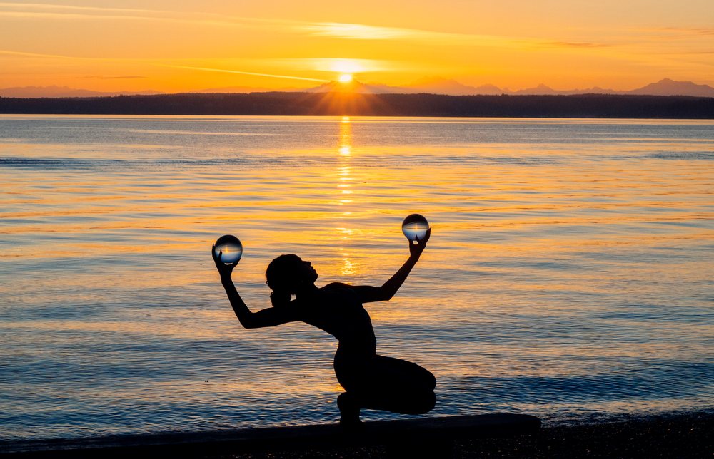 Silhouette of a woman performing a yoga acrobatic balancing pose with crystal balls at sunset over water, USA.