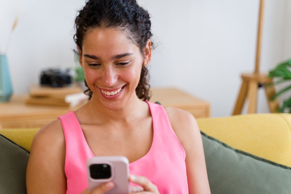 A smiling woman wearing a pink top is using her smartphone while sitting on a yellow sofa in a modern living room, Spain.