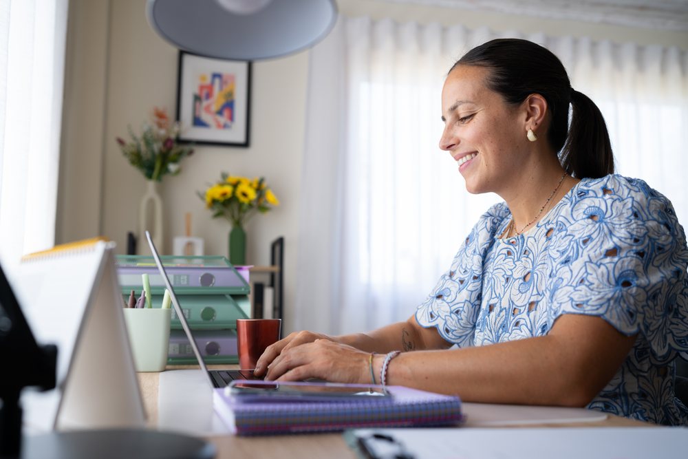 Woman typing on a laptop at a desk in a home office with sunlight through a window, telecommuting, Spain.