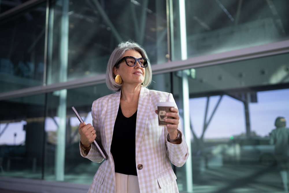 Businesswoman wearing glasses and plaid blazer holding a coffee cup and tablet outside a modern office building, Spain, Madrid.