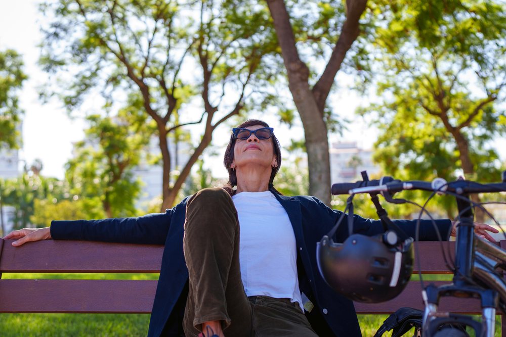 Woman relaxing on a bench in a park with her bicycle and helmet beside her under trees in sunlight, Málaga.