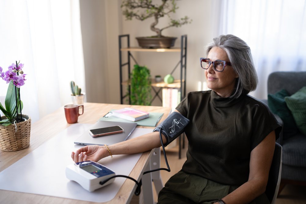 Woman with gray hair and glasses measures blood pressure using a digital monitor while sitting at a table in a well-lit home interior with plants and a laptop, Spain.