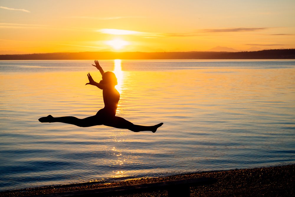 Silhouette of a woman performing a split leap yoga pose at sunset by the water, USA.