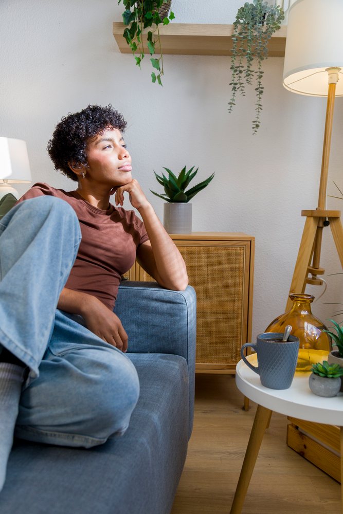 Young woman sits thoughtfully on a grey sofa in a modern living room with potted plants and a coffee mug on a white round table nearby.