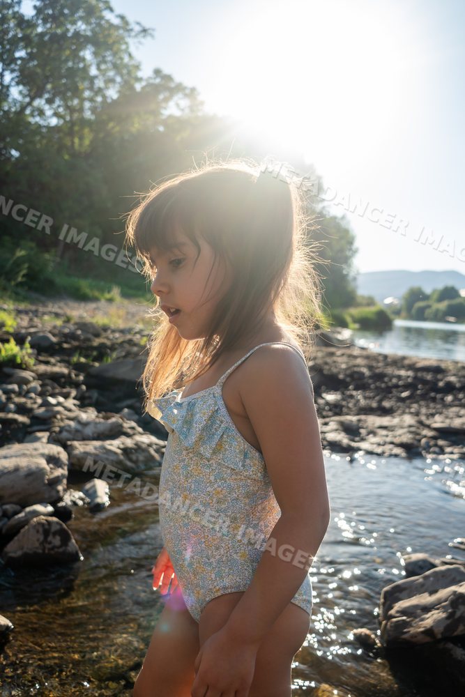 A white girl in a swimsuit explores rocky riverbank water on a sunny