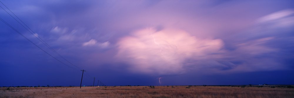 Lightning flashes from a thunderhead cloud over a dry grass field with power lines at dusk.