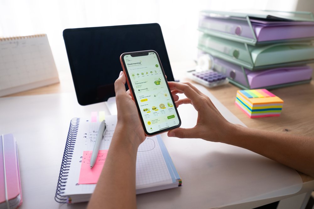 Hands holding smartphone showing food delivery app over desk with notebook and office supplies at home, Spain.